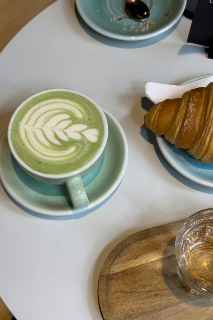 Matcha latte with leaf-pattern latte art in a teal cup served beside a croissant