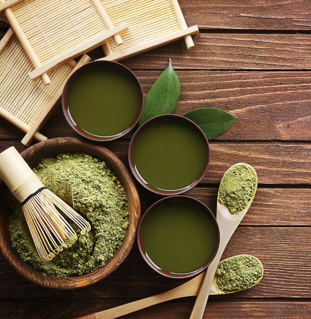 Three bowls of matcha tea with a bamboo whisk, tea leaves, and matcha powder on wooden spoons