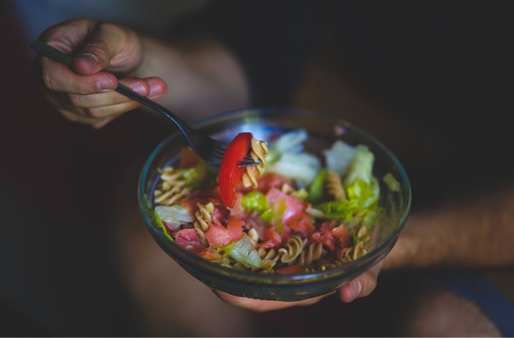 Person eating a pasta salad with vegetables from a glass bowl with a fork