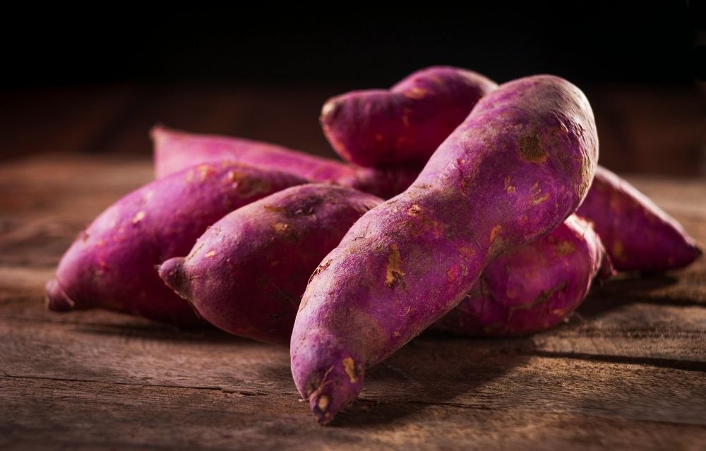 Whole raw purple sweet potatoes with deep violet skin and flesh on a dark wooden surface