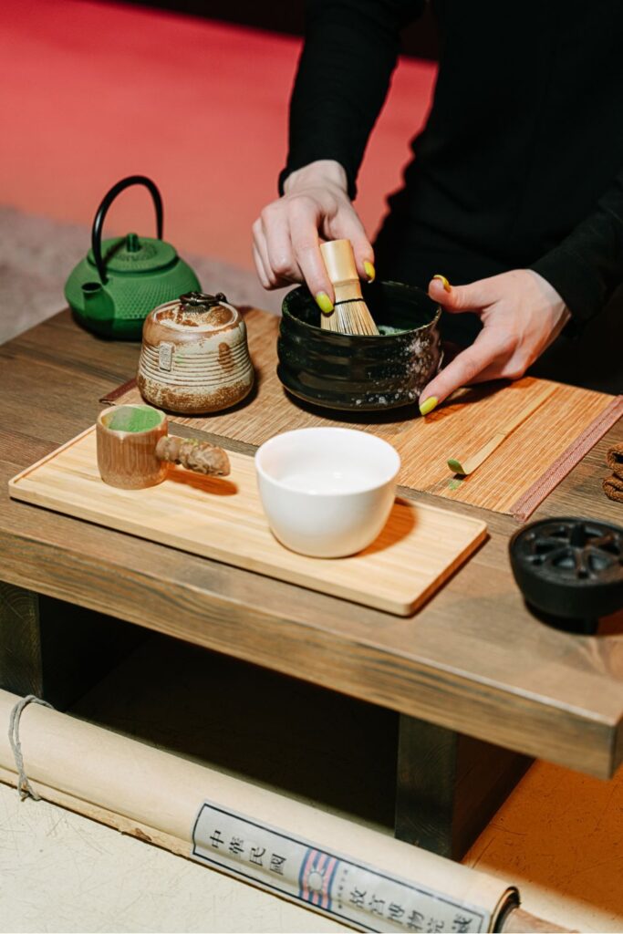 Person preparing matcha tea with a bamboo whisk in a black bowl, with a green teapot and cups nearby