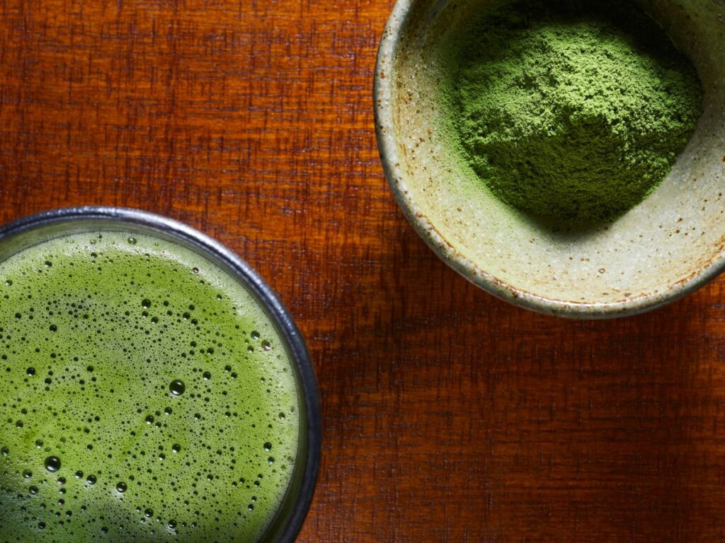 Frothy Matcha drink next to a bowl of Matcha powder on a wooden table