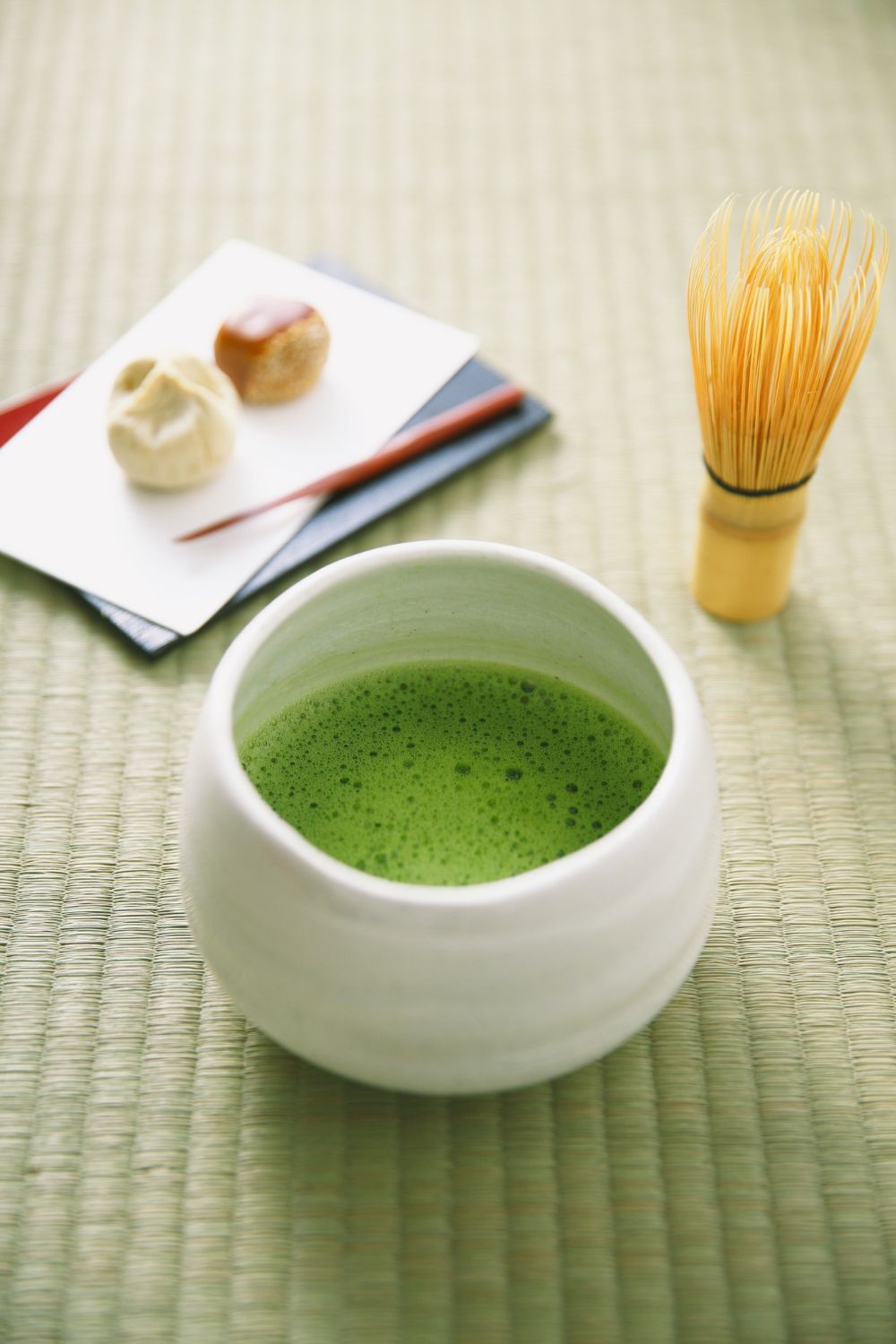 A bowl of frothy matcha tea with a bamboo whisk beside a small plate of Japanese sweets on a tatami mat