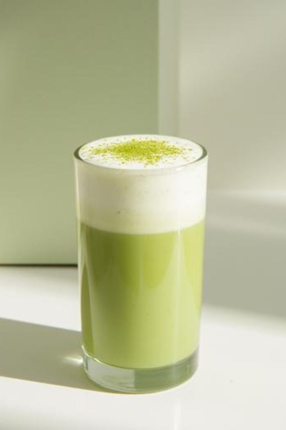 A minimalist matcha latte in a tall glass with milk foam on top dusted with matcha powder, against a plain green background