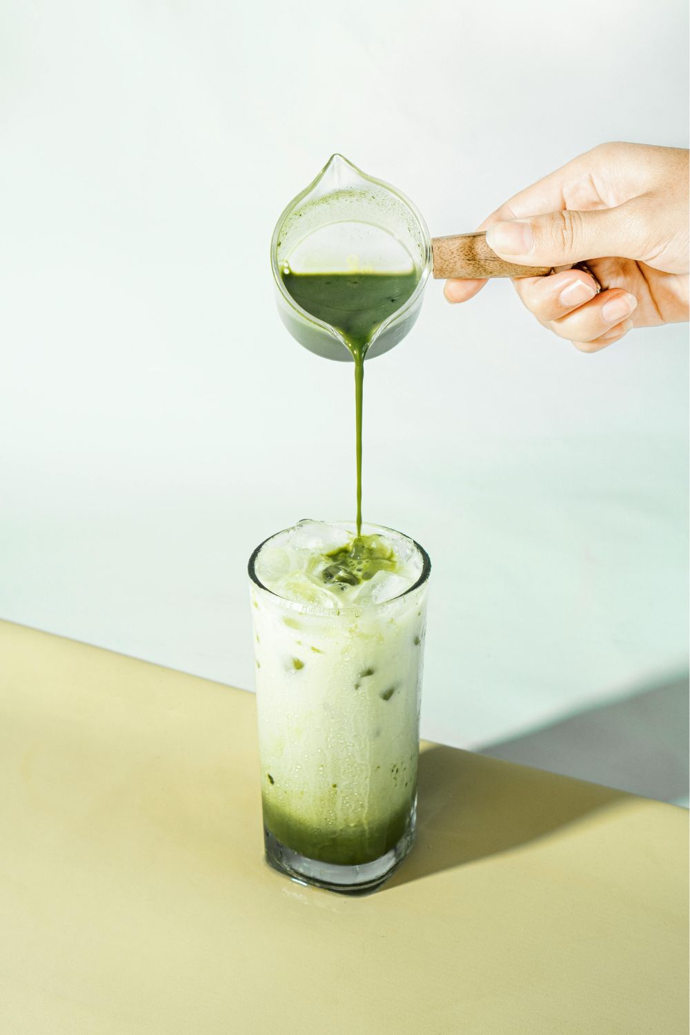 A hand pouring green matcha syrup from a small glass pitcher into a tall glass of iced latte with milk