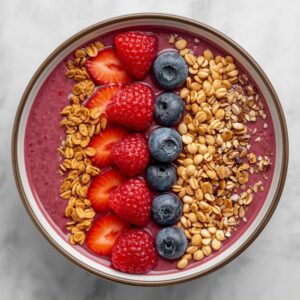A bowl of berry smoothie topped with sliced strawberries, raspberries, blueberries, and granola
