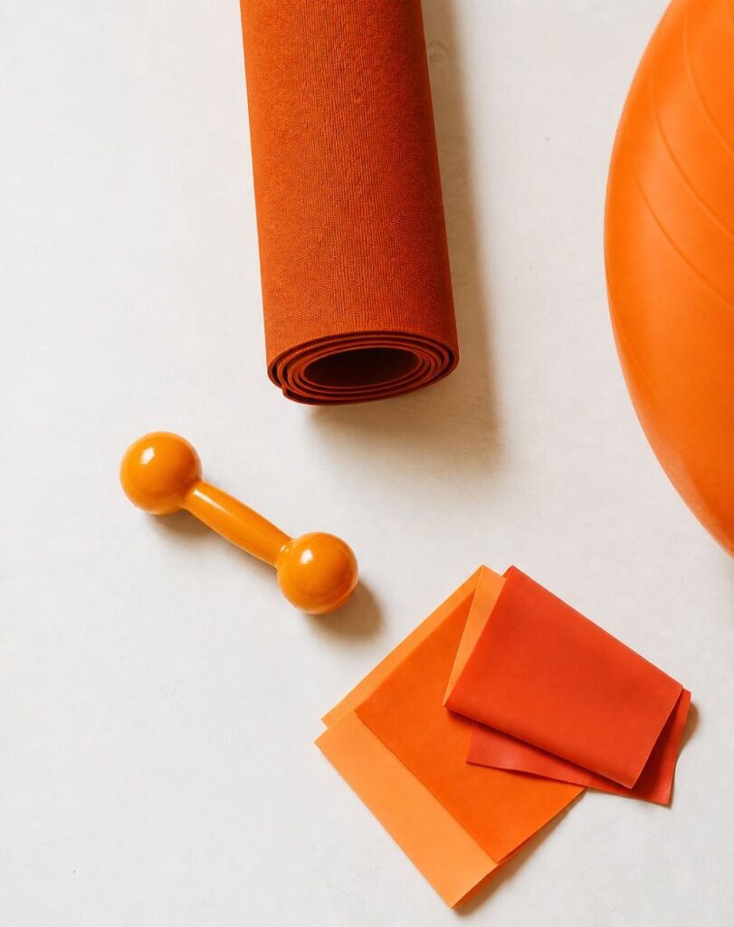 Flat lay of exercise equipment including a rolled yoga mat, dumbbell, and resistance bands in orange tones