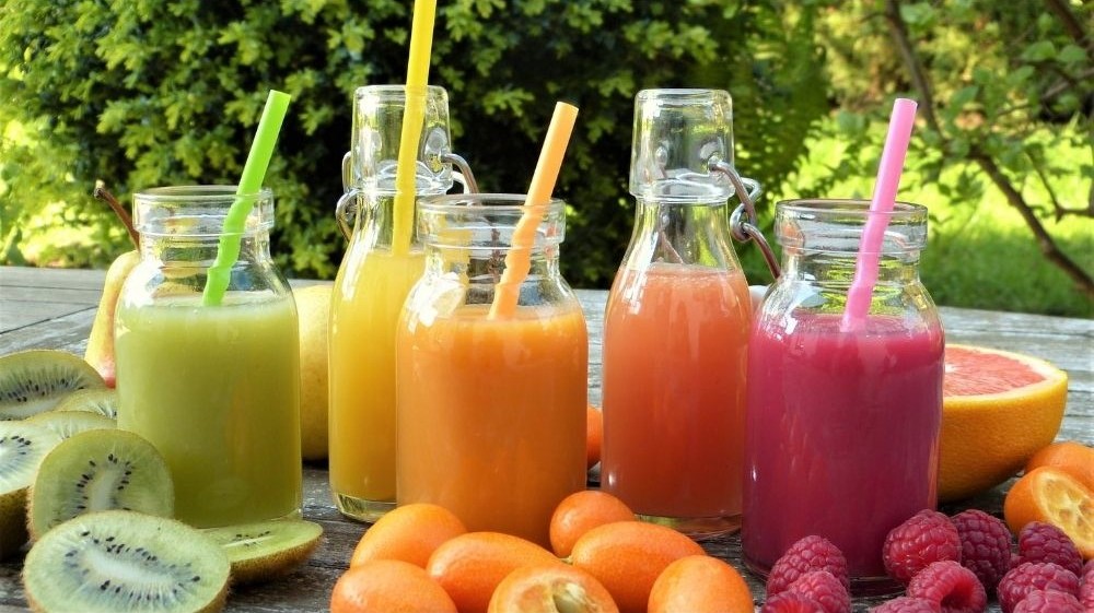 Five colorful smoothies in glass bottles with straws, surrounded by kiwi, citrus fruit, and berries